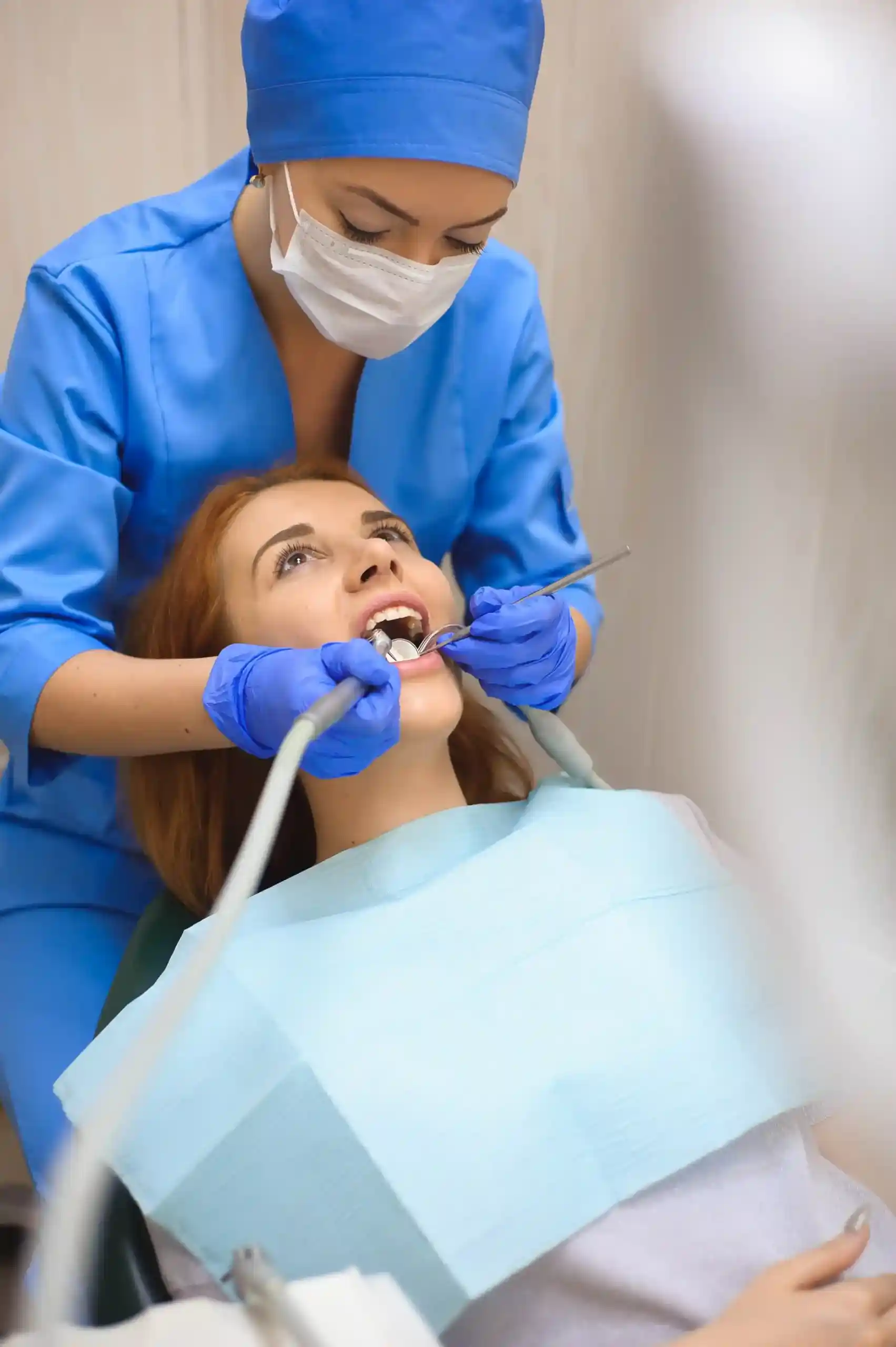 A dentist in blue scrubs and a face mask performs an oral surgery procedure on a patient, using a dental handpiece and a mirror to work inside the patient's mouth.