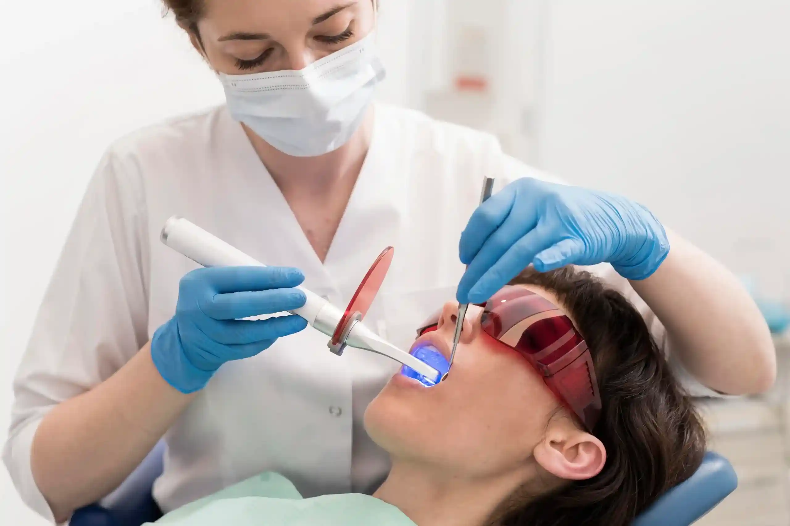 A dentist uses a blue dental curing light or laser on a patient wearing protective red goggles during a clinical procedure.